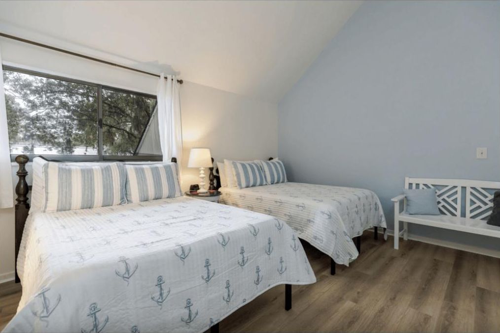 Un dormitorio con dos camas, una ventana con cortinas, una mesita de noche con lámpara y un banco.