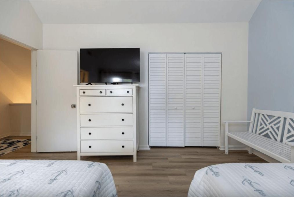Une chambre avec deux lits, une commode avec un téléviseur, une armoire blanche et un banc.