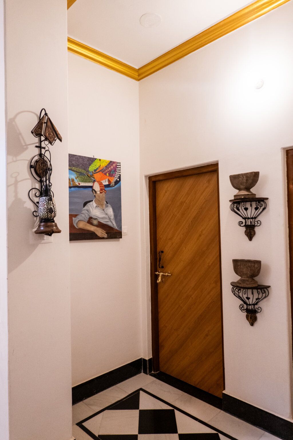 A hallway with a wooden door, a black and white checkered floor, a wall-mounted light fixture, and a colorful painting.