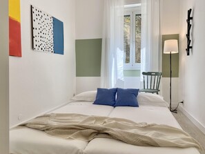 A bedroom with a bed, pillows, a blanket, a chair, a floor lamp, and a window with curtains.
