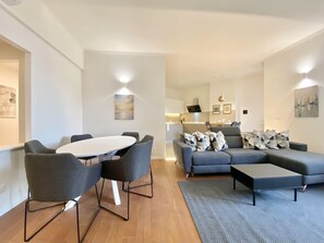 Una sala moderna con un sofá gris, una mesa de comedor redonda con sillas, y una cocina al fondo.