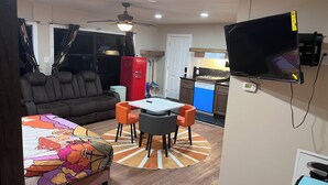 A compact living space with a sofa, a small dining table with chairs, a red refrigerator, a TV mounted on the wall, and a kitchen area with a dishwasher.