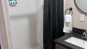 A bathroom with a shower, white tiled walls, a black shower curtain, a white towel on a ring, and a wooden cabinet.