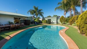 Ein Pool mit geschwungener Kante, umgeben von einer Ziegelkante und künstlichem Rasen. Im Hintergrund sind Palmen und weitere Grünpflanzen zu sehen.