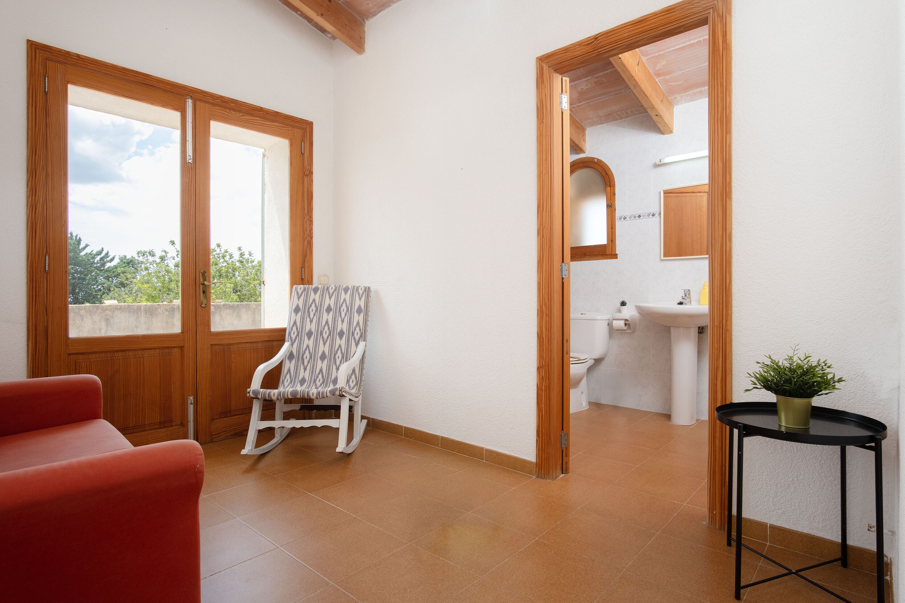 A room with a wooden door, a rocking chair, a red sofa, a small table with a plant, and a bathroom with a sink and mirror.