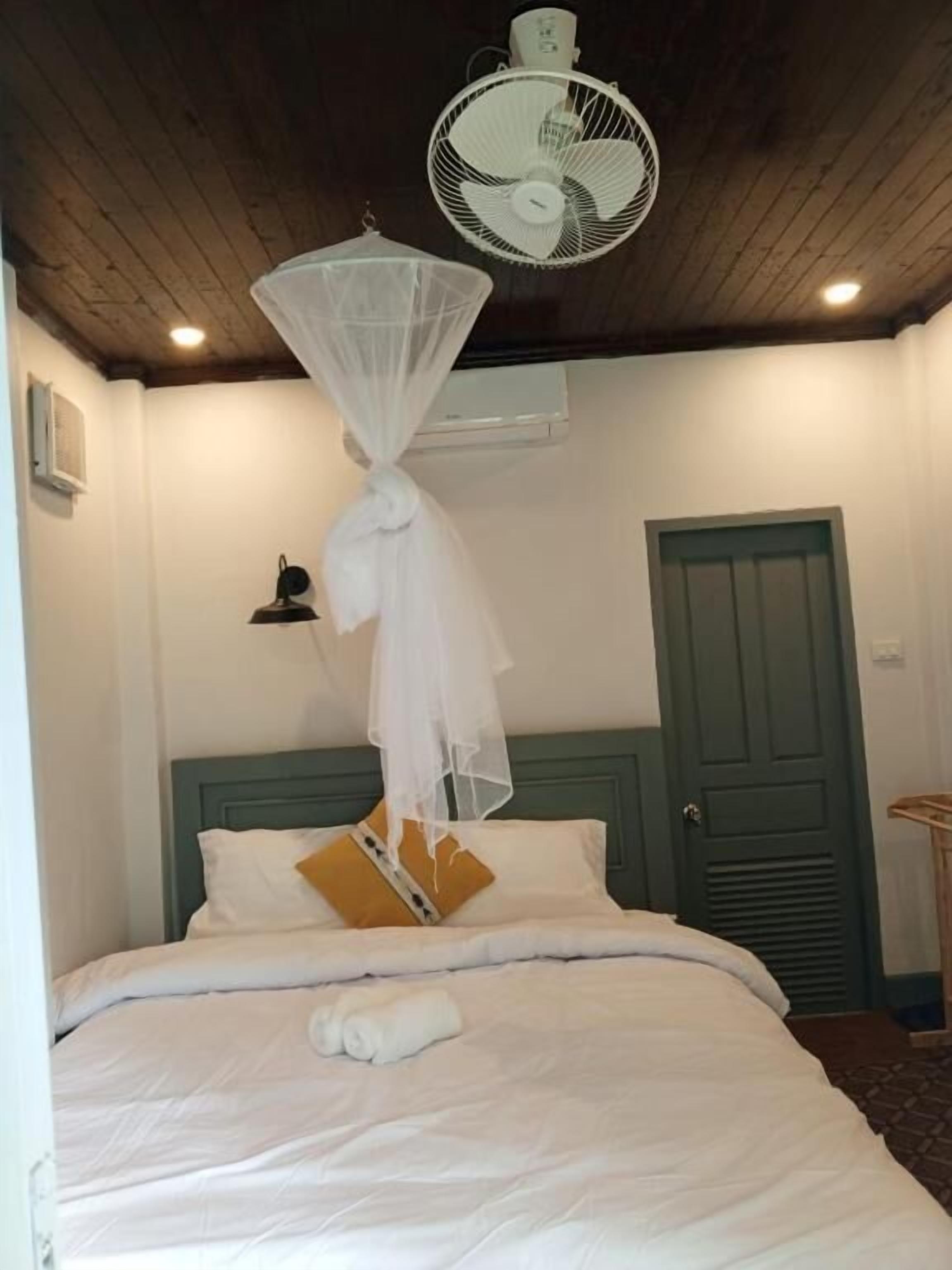 Une chambre à coucher avec un lit, du linge de lit blanc, un baldaquin blanc, un ventilateur de plafond, une porte verte et une petite table avec une lampe.