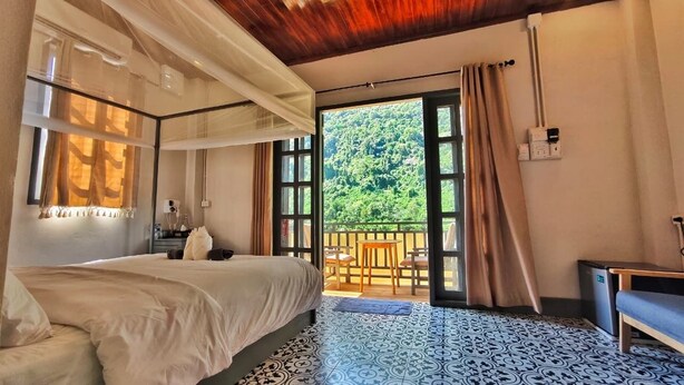 A bedroom with a bed, a balcony with a table and chairs, and a view of greenery outside. - Dokchampa River View Villa (Nong Khiaw)