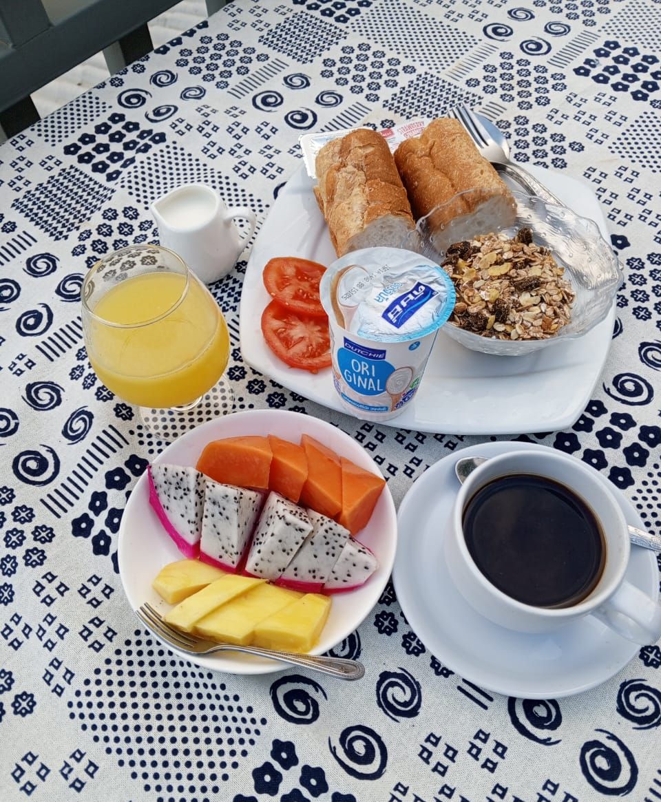 Pöydällä on kuvioitu pöytäliina, lasi appelsiinimehua, lautanen, jossa on leipää, tomaatteja, jogurttia ja mysliä, sekä kuppi mustaa kahvia.