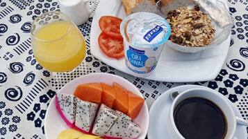 Pöydällä on kuvioitu pöytäliina, lasi appelsiinimehua, lautanen, jossa on leipää, tomaatteja, jogurttia ja mysliä, sekä kuppi mustaa kahvia.