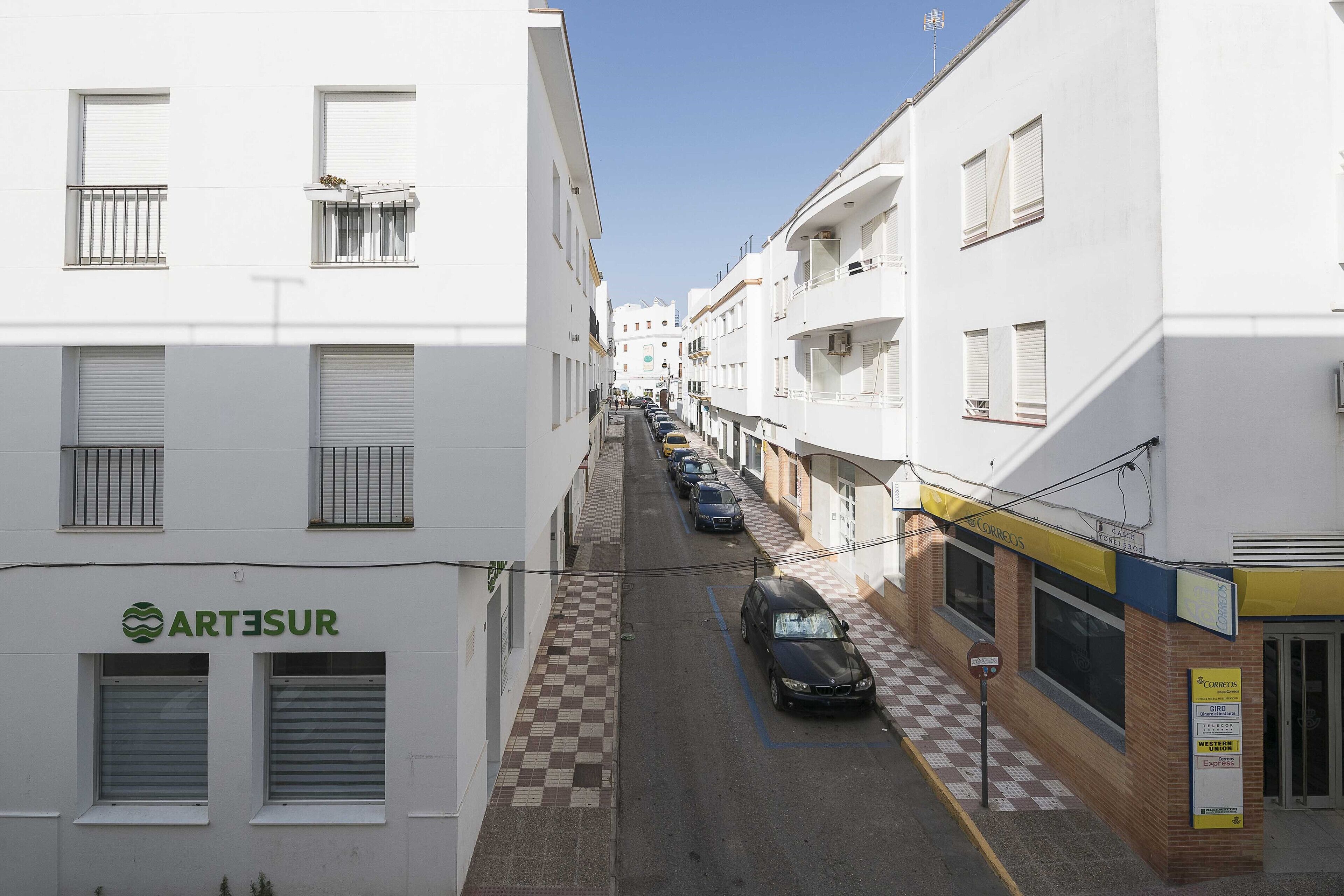 Une rue étroite bordée de bâtiments blancs, une voiture garée et une devanture de magasin bien visible, avec un auvent jaune.
