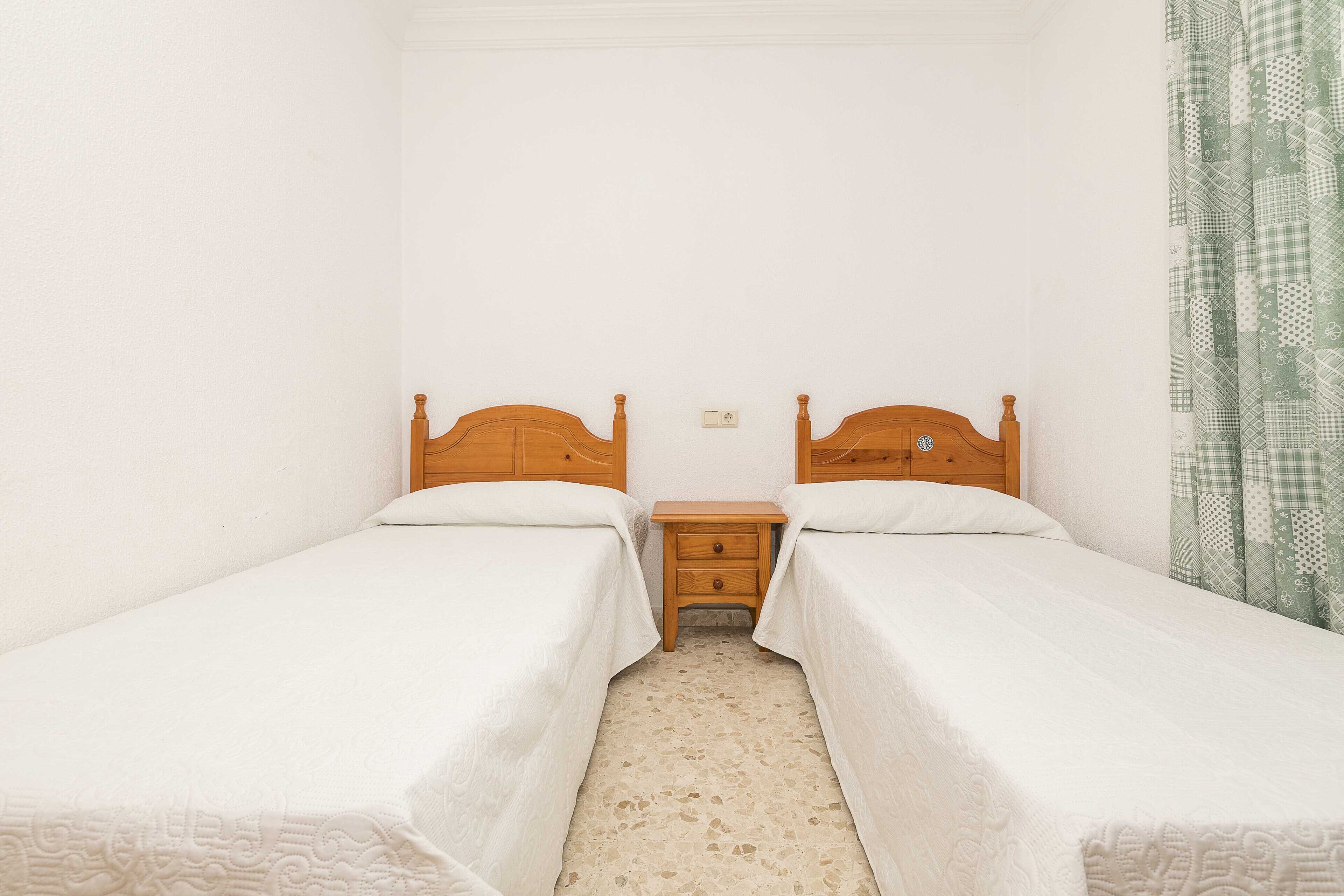 Une chambre avec deux lits simples, une table de chevet en bois placée entre les deux, et un rideau à motifs sur le côté droit.