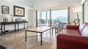 A spacious living area with a red sofa, a wooden coffee table, and a dining table. There are framed pictures on the wall, a lamp, and a view of the sea through large windows. - ES TAMARELLS (S'ESTANYOL) - Villa by the Sea with Private Pool and Free Wi‑Fi (Artà)