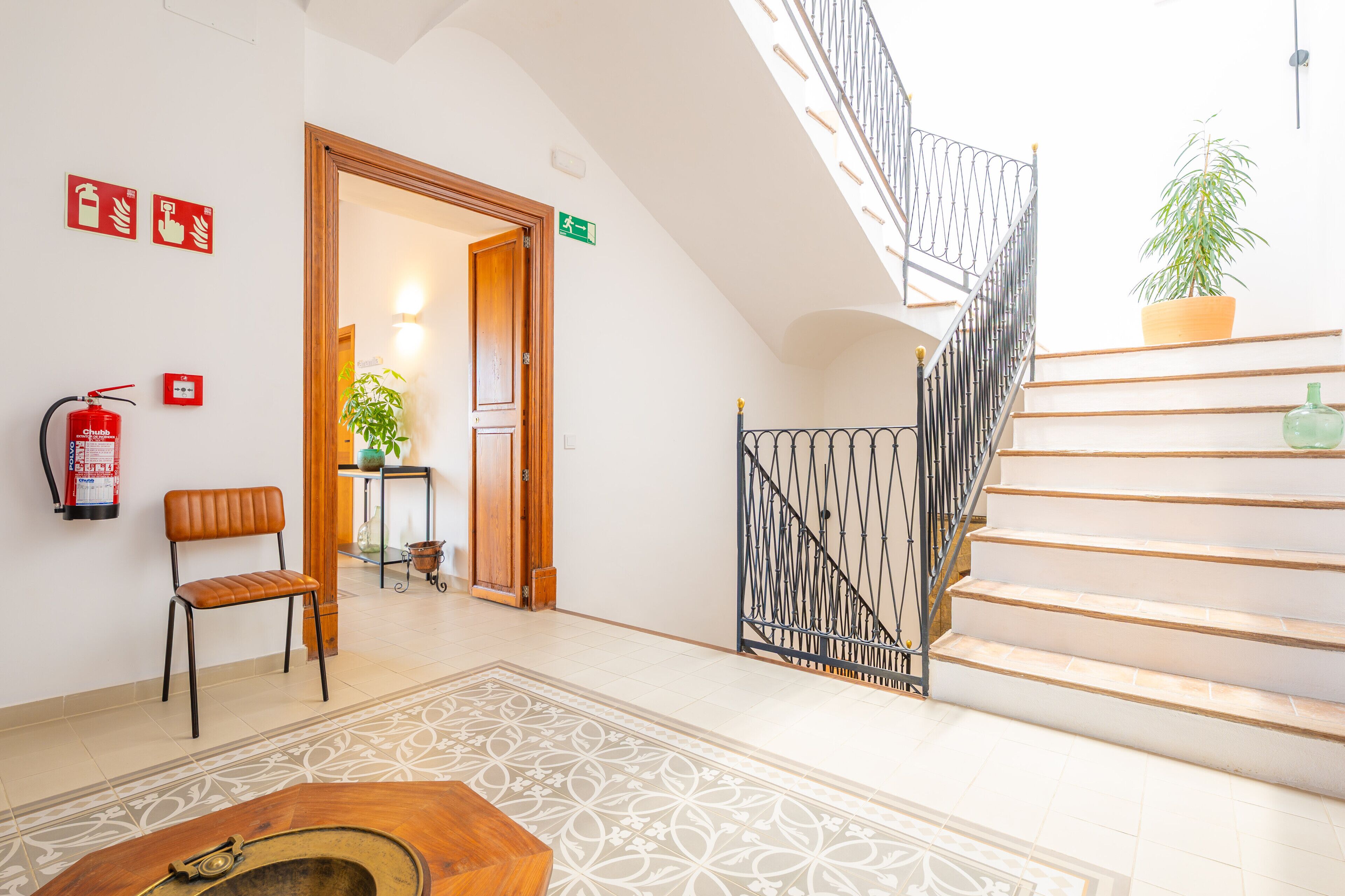 A hotel lobby with a staircase, a fire extinguisher, a chair, a plant, and a patterned rug.