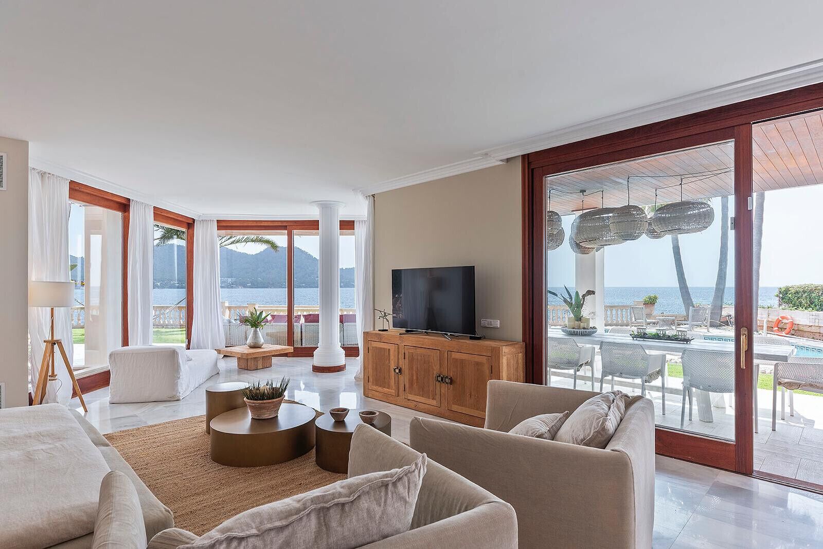 A spacious living room with a large TV, comfortable seating, a wooden cabinet, and a view of the sea through large sliding doors.