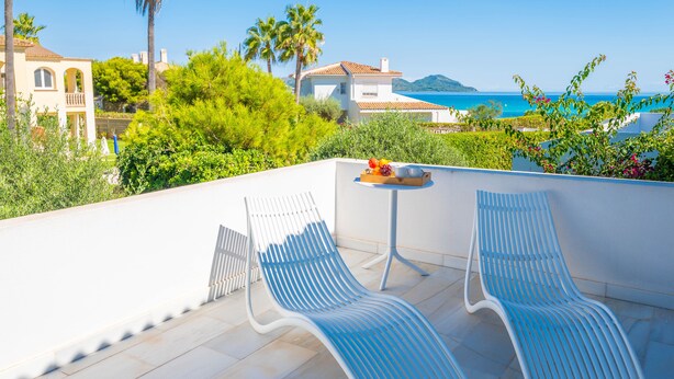 Une terrasse avec deux fauteuils blancs, une petite table sur laquelle sont disposés des fruits, et une vue sur la mer et les bâtiments.