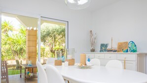 Un coin repas avec une table et des chaises blanches, une table à plateau en verre, et une vue sur un jardin grâce à de grandes fenêtres.