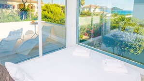 Une chambre avec une grande fenêtre donnant sur un jardin et la mer. On y trouve deux chaises blanches et une petite table sur laquelle est posée une serviette.