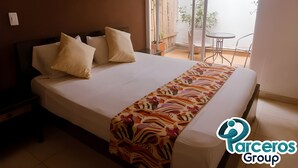 A hotel room with a large bed, two pillows, a colorful bedspread, a nightstand, a small table, and a view of the outside balcony. - PARCEROS GROUP-VACATION ROOM FOR COUPLES, FLOOR 1, PEREIRA (pereira)