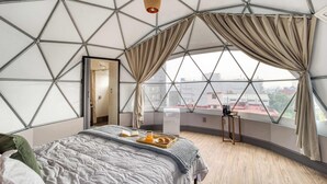 Ett rum med ett geometriskt mönstrat tak, en säng med ett brickbord med frukost och utsikt över staden genom stora fönster.