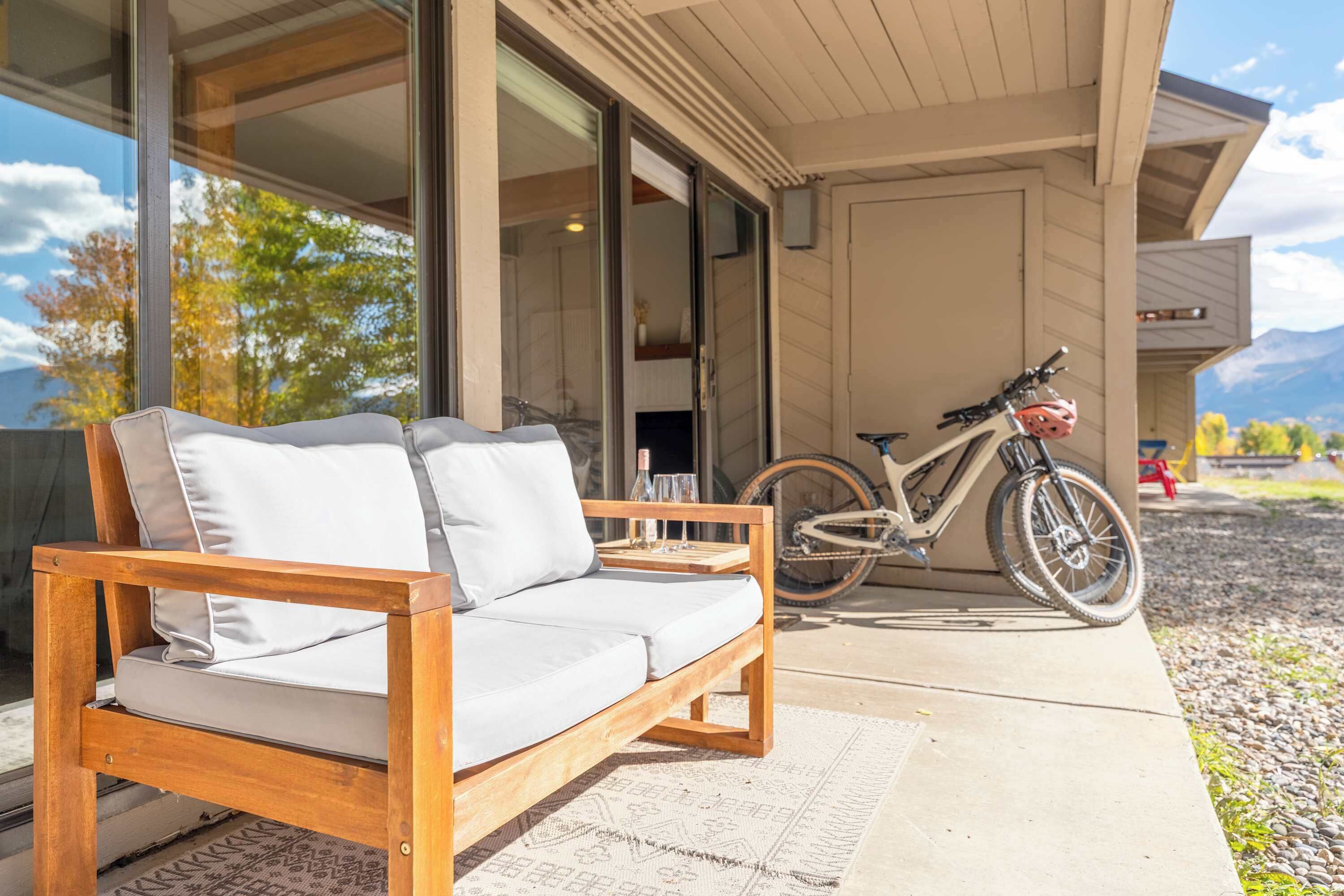 A wooden sofa with cushions on a patio, a bicycle, and a view of mountains through a glass door.