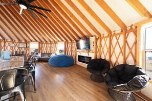 A spacious room with a high ceiling, wooden beams, a dining area with chairs, a blue bean bag, and a TV on a wall. - OsoBlu Yurts Group Booking (Garden City)