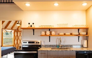 A kitchen with a stove, oven, sink, and shelves with glassware and plates. - OsoBlu Yurts Group Booking (Garden City)