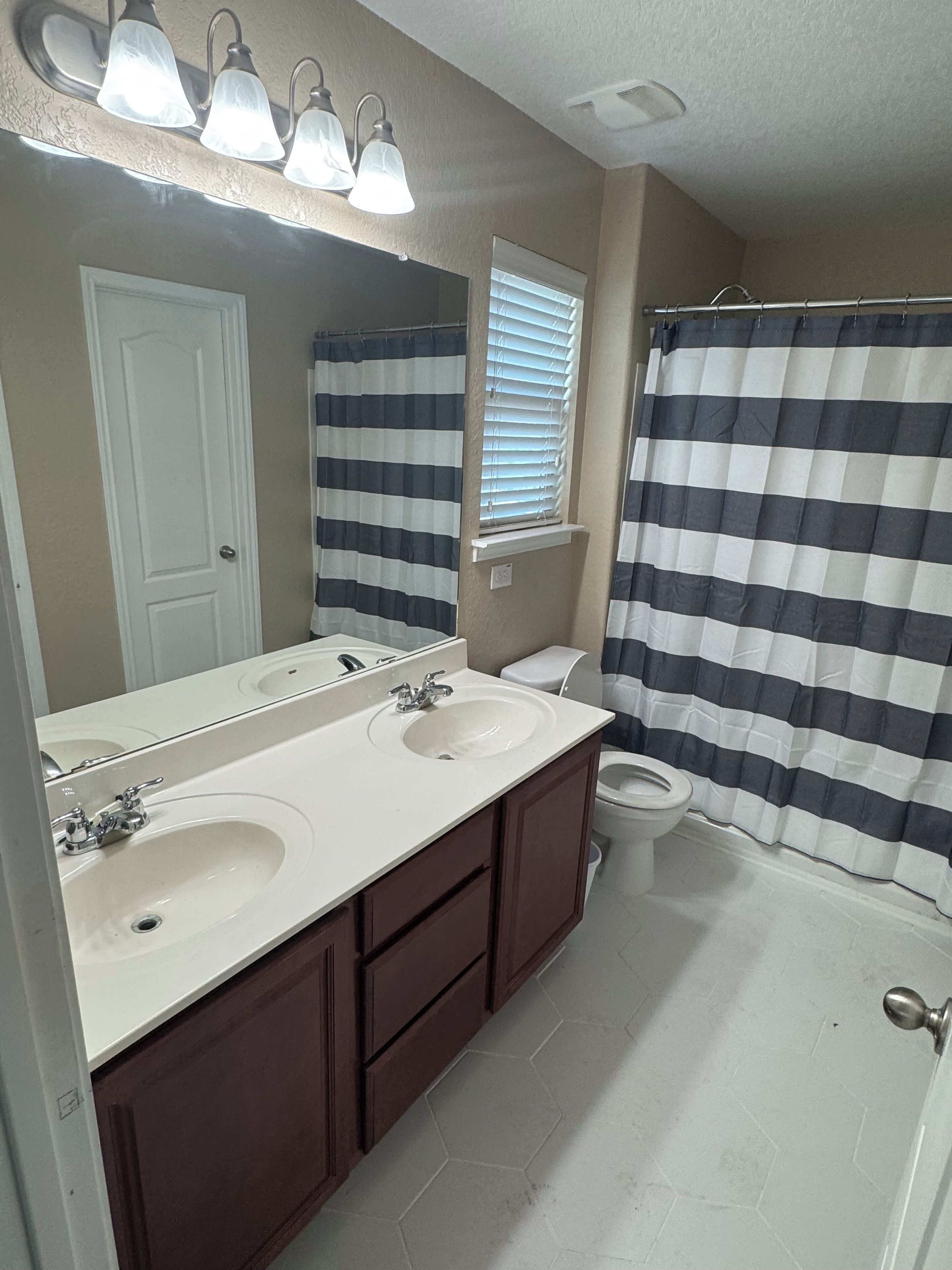 A bathroom with two sinks, a toilet, a shower curtain, and a window with blinds.