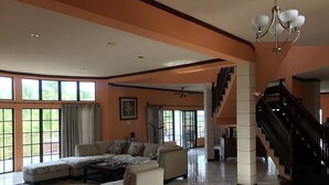 Ein geräumiges Wohnzimmer mit einem Ecksofa, einem Couchtisch und einem gemusterten Teppich. Eine Treppe führt auf eine höhere Ebene, und große Fenster sorgen für natürliches Licht.