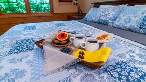 Cama bem arrumada com colcha azul e branca, bandeja com itens de café da manhã incluindo pão, frutas e café, e vista para o verde através de uma janela.