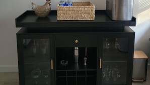 Eine Küchenarbeitsplatte mit einem gewebten Korb, der Wasserflaschen, einen Edelstahlkessel und eine dekorative Hähnchenfigur enthält. Darunter befinden sich zwei schwarze Schränke mit Glastüren, einer mit einer Kaffeemaschine und der andere mit Gläsern.