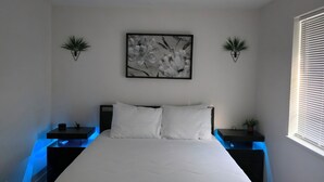 Ein Schlafzimmer mit einem Bett, zwei Nachttischen, einem Wandbild und einem Fenster mit Jalousien.