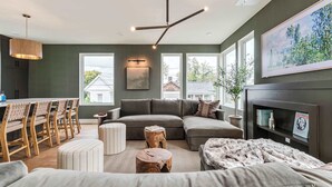 Uma sala de estar moderna com sofá seccional, uma área de jantar com cadeiras de madeira, uma luminária de teto diferenciada e uma lareira com livros e objetos decorativos.