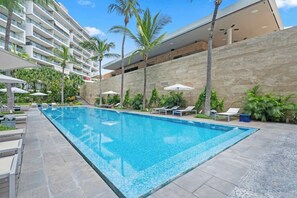 A swimming pool with lounge chairs and umbrellas, surrounded by palm trees and a stone wall.