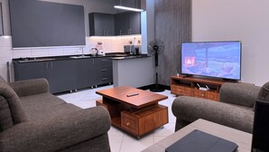 Un salon moderne doté d’un téléviseur à écran plat, de canapés gris, d’une table basse en bois et d’un coin cuisine avec des meubles sombres et un plan de travail blanc.