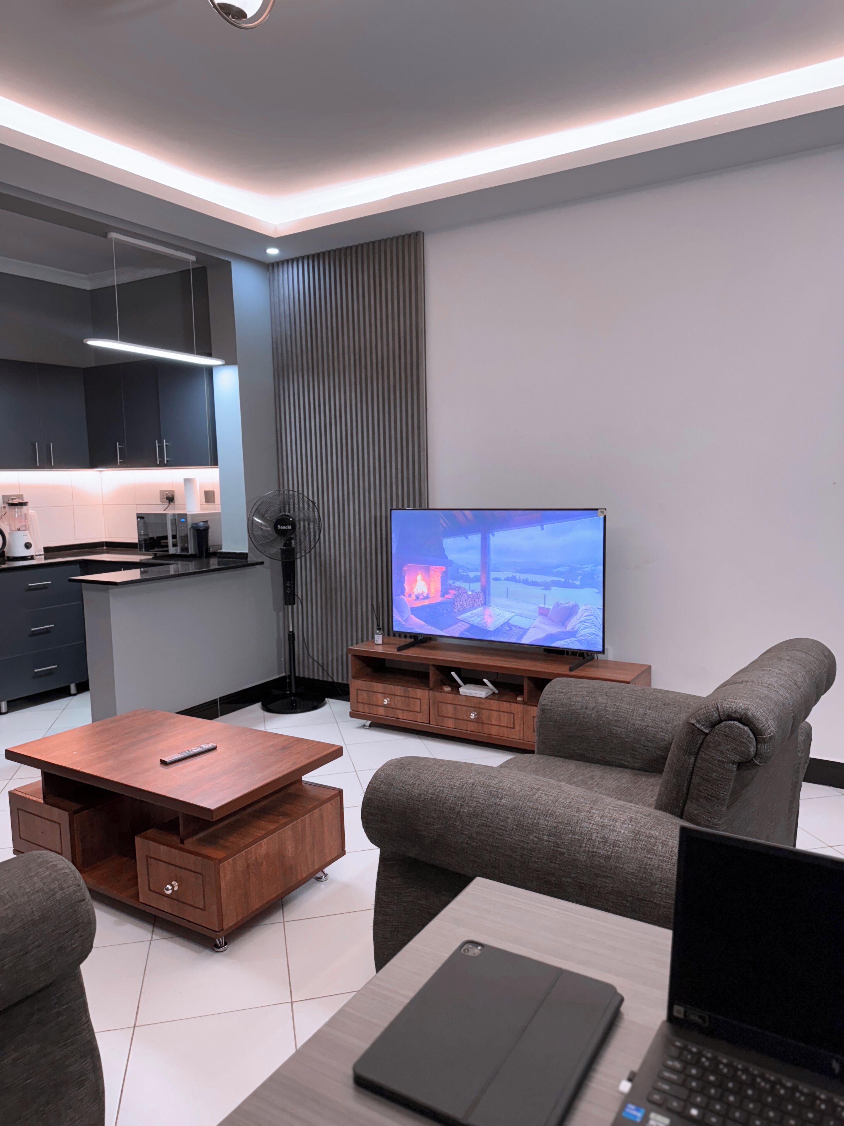 A modern living room with a flat-screen TV, grey sofas, a wooden coffee table, and a laptop on a small table.