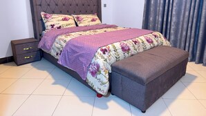 Un lit doté d’une tête de lit capitonnée, d’un pied de lit et d’un banc. On y trouve une table de chevet avec des tiroirs et une lampe.