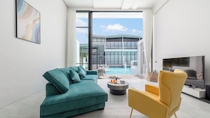 A modern living room with a teal sofa, a yellow armchair, a flat-screen TV, and a view of a pool and building through large windows. - Unique Townhouse Pool Slide Access (Bophut)