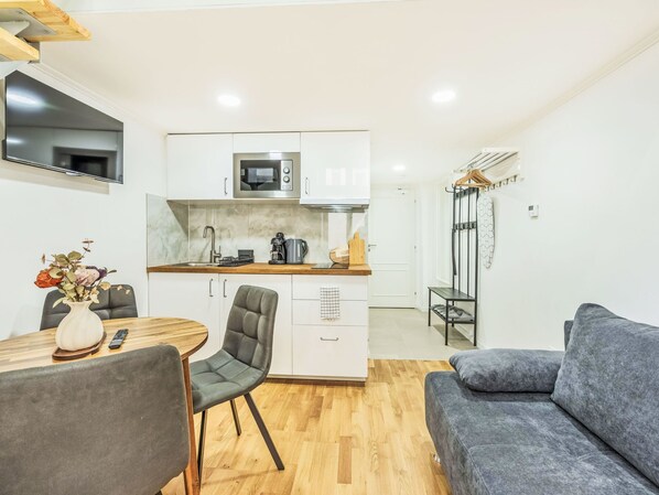 Un espacio habitable compacto con kitchenette, una zona de comedor con mesa y sillas, y un sofá gris.