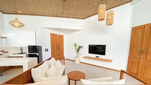 A modern living room with a wooden sectional sofa, a TV mounted on the wall, and a kitchen in the background. - Maravilla two bedrooms with garden view (Bali)