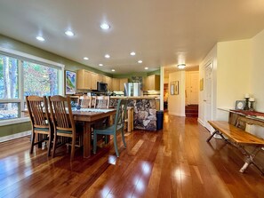 A spacious kitchen with wooden cabinets, a dining area with a wooden table and chairs, and a view of trees through large windows.