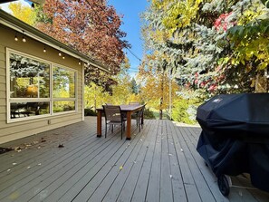 A wooden deck with a table and chairs, a barbecue grill, and a house with large windows.