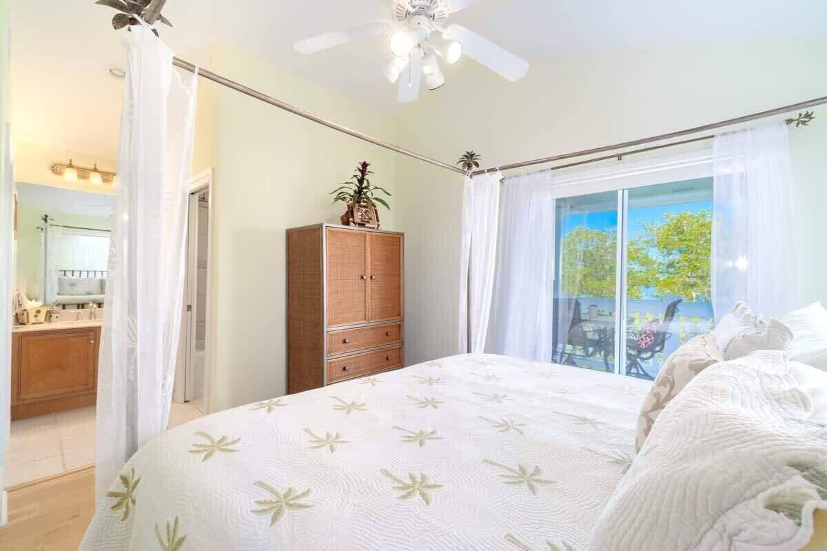 A bedroom with a large bed, a wooden cabinet, a ceiling fan, and a window with a view of trees.