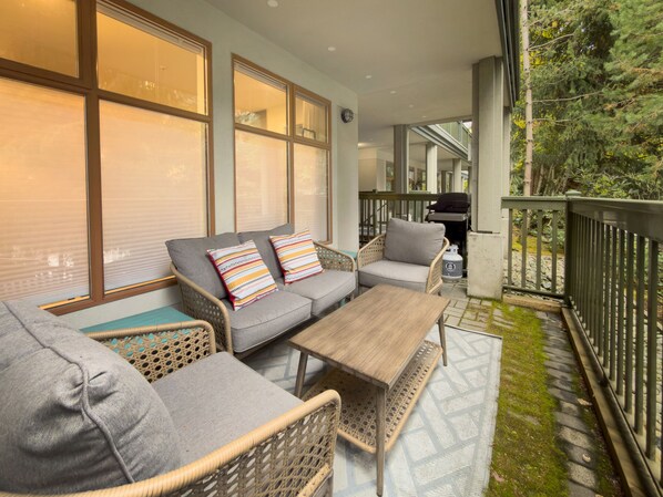 A covered outdoor seating area with a sofa, chairs, and a table, surrounded by large windows and a railing.
