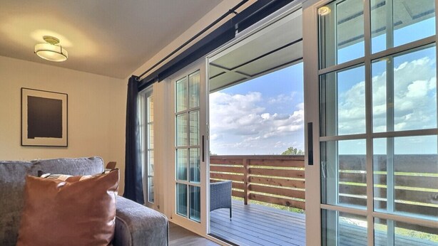 A room with a grey sofa, a brown pillow, and a large sliding glass door leading to an outdoor deck with a wooden railing. - City Overlook w/ Pool & Extra Large Balcony (Birmingham)
