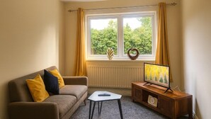 Un salon avec un canapé, une petite table, un téléviseur posé sur un meuble en bois et une fenêtre avec des rideaux.