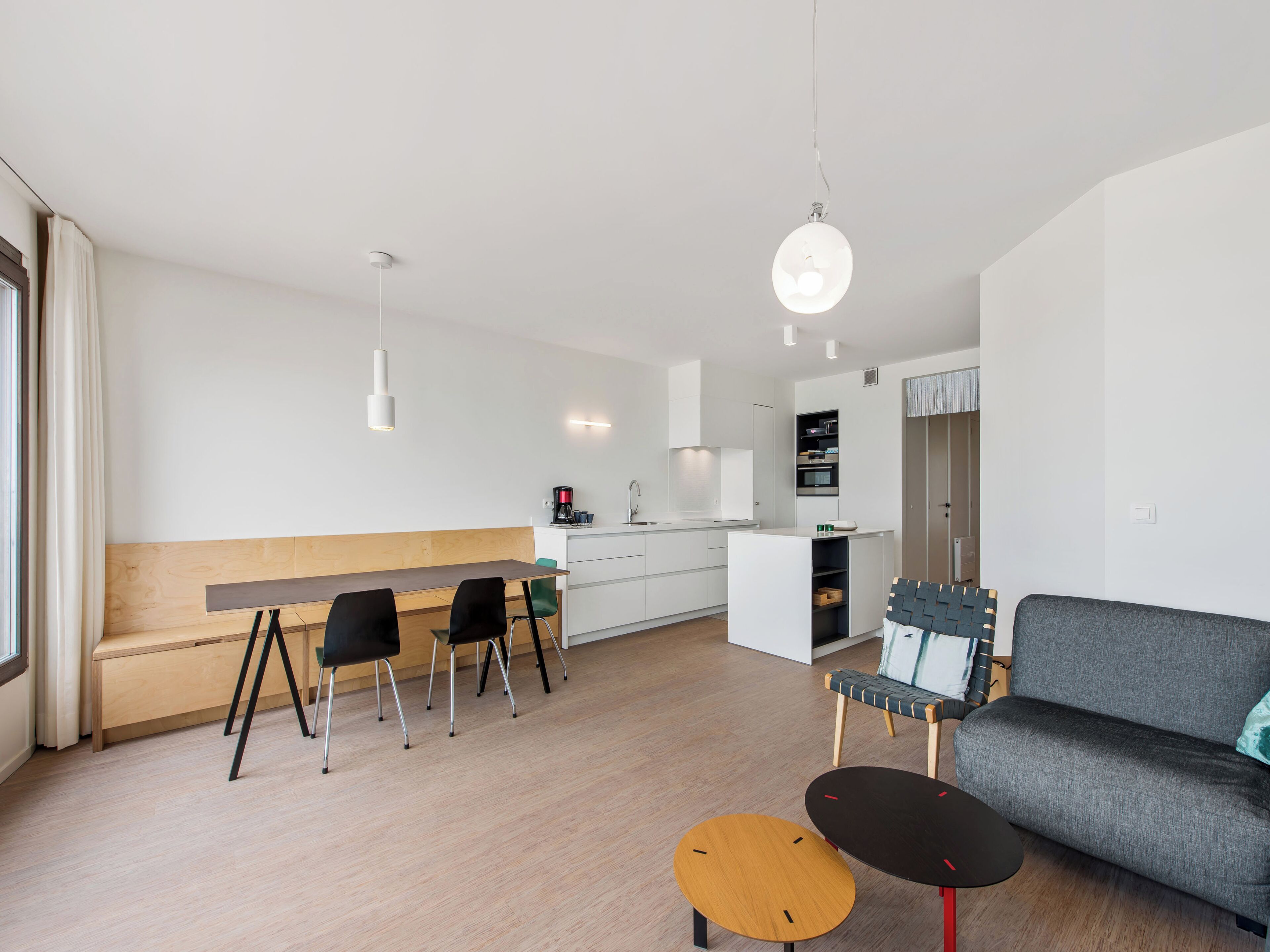 A modern, minimalist living and dining area with a wooden bench, black chairs, a grey sofa, and a small round table.