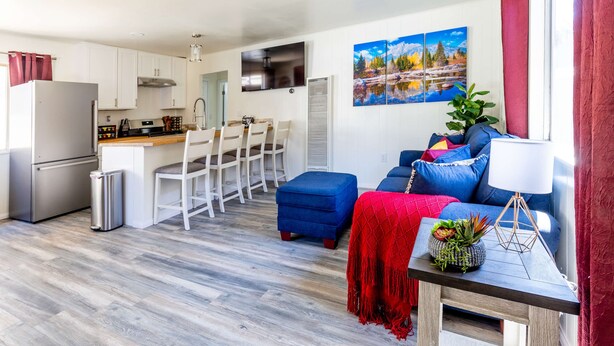 Une cuisine moderne avec des armoires blanches et des appareils en acier inoxydable, un coin repas avec des chaises blanches et un espace de vie avec un canapé bleu et un plaid rouge.