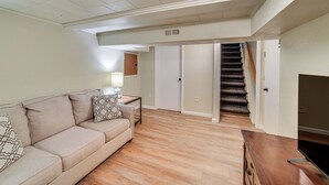 Una sala de estar con un sofá color beige, una mesa de centro de madera y un mueble para televisor. Se ve una escalera que sube a un nivel superior.