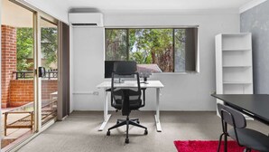 Un espace de bureau moderne comprenant un bureau, une chaise et un ordinateur, de grandes fenêtres donnant sur un paysage verdoyant, et un tapis rouge.
