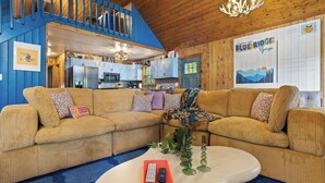 A cozy living room with a large beige sectional sofa, a white coffee table, and a kitchen in the background.